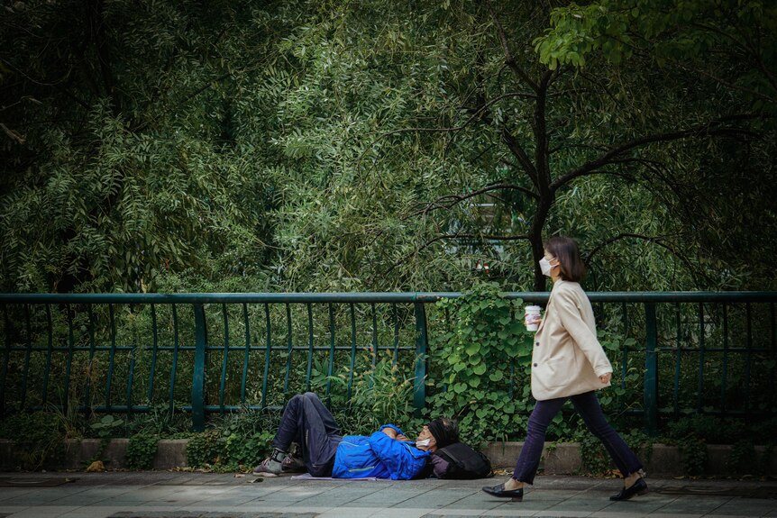 A woman wearing a coat and masks walks by a homeless man laying on the ground.