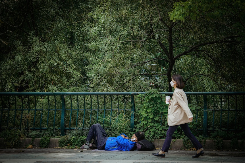 A woman wearing a coat and masks walks by a homeless man laying on the ground.