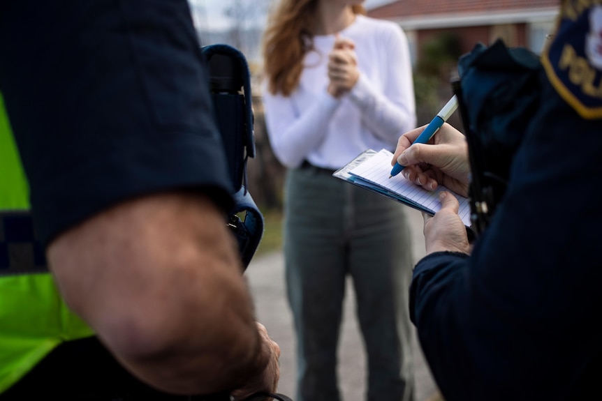 A police officer writes in a notebook as a young woman stands in the background