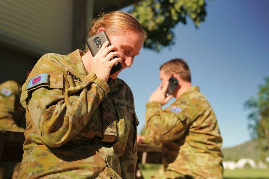An army corporal sits on a seat outside in uniform, talking on the phone and smiling.