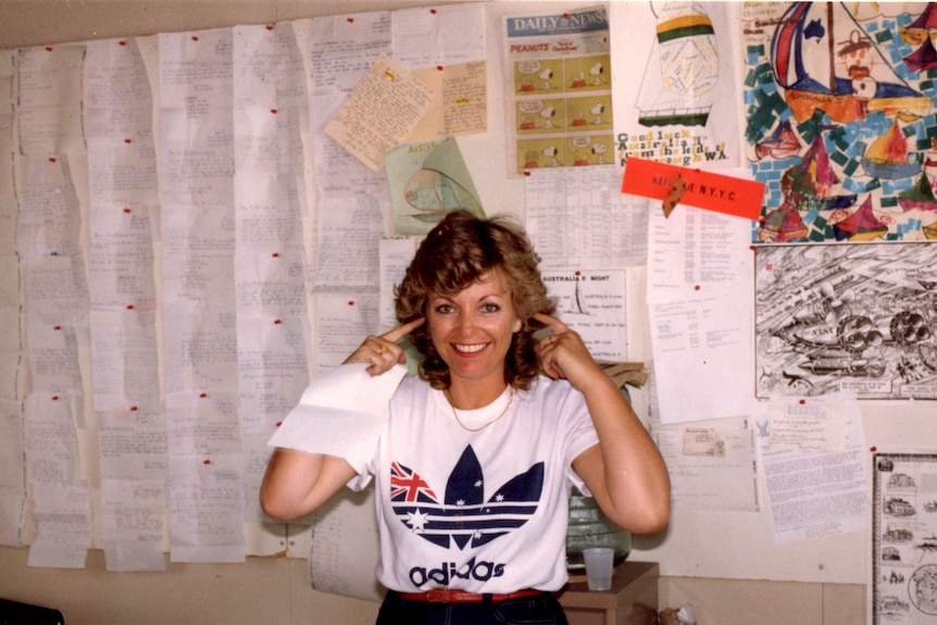 A smiling woman stands blocking her ears in an office with numerous documents on the wall.