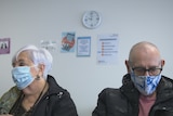 A woman and man wearing masks sit in the waiting room of a GP clinic. On the wall behind them is a clock.