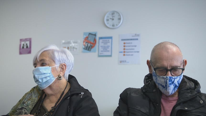 A woman and man wearing masks sit in the waiting room of a GP clinic. On the wall behind them is a clock.