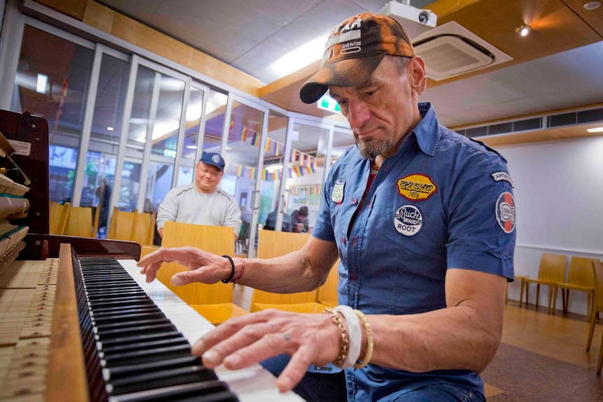 A man in a blue shirt and camouflage cap plays the piano