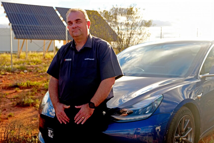 A man leans on an electric car in front of a solar panel. 