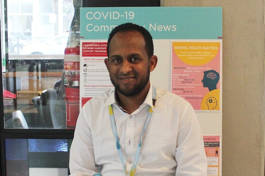 A man wearing a white shirt and a lanyard sits in a foyer with a coronavirus information board behind him.