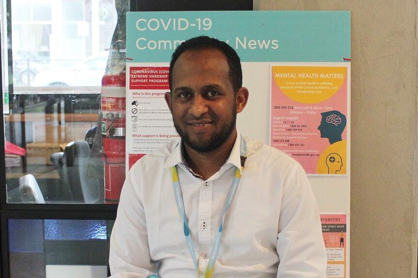 A man wearing a white shirt and a lanyard sits in a foyer with a coronavirus information board behind him.