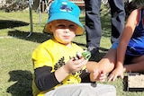 A young boy in a blue shirt plays with toy while his mouth is full.