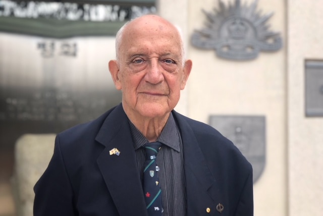 Colin Kahn looks directly at the camera. He wears a navy blazer, dark shirt and blue and green tie. He has medals on his lapel.