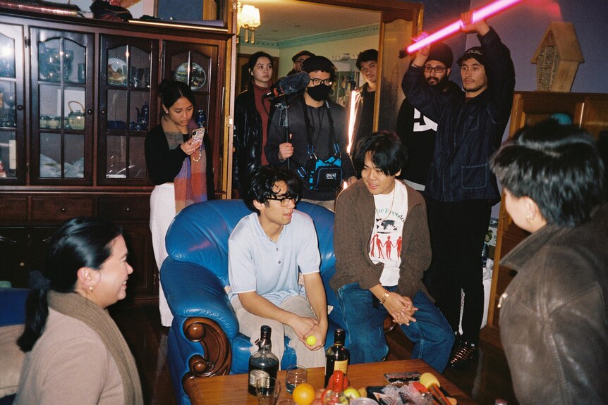A film set inside a house with two men sitting on a couch surrounded by crew.