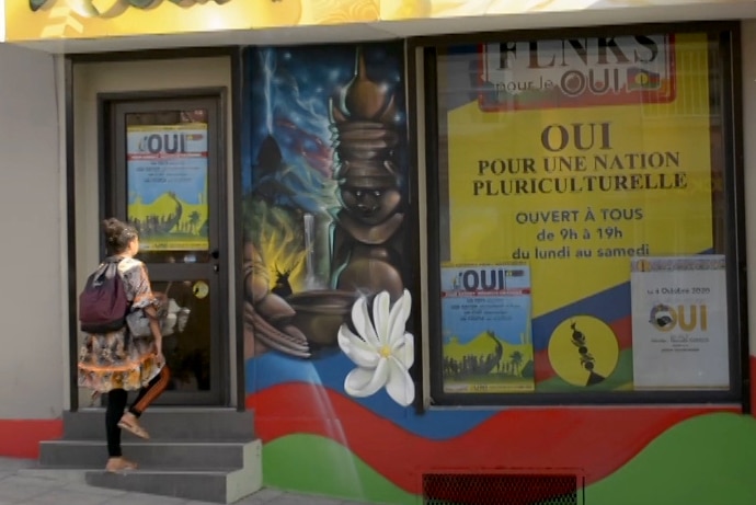 A woman walks up steps to the doors of a glass-fronted building covered with bright colours of New Caledonia's Kanak flag.