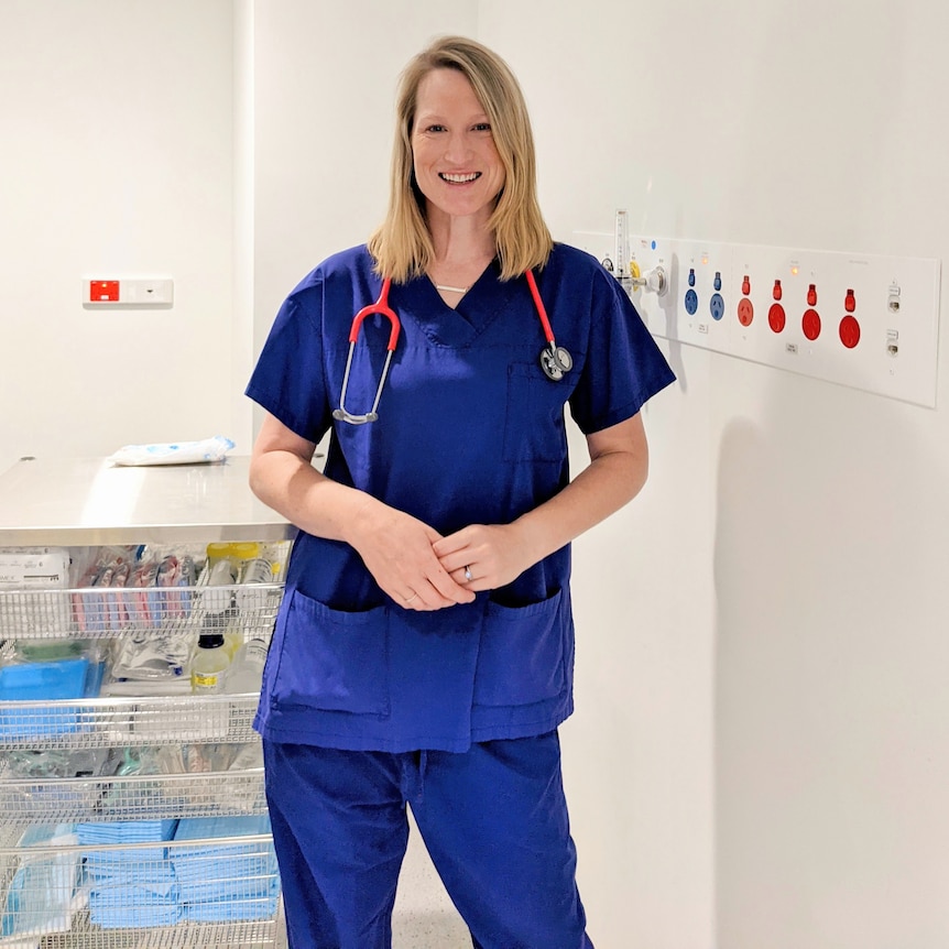 A young woman with straight blonde hair, a blue medical uniform, and a stethoscope, smiles