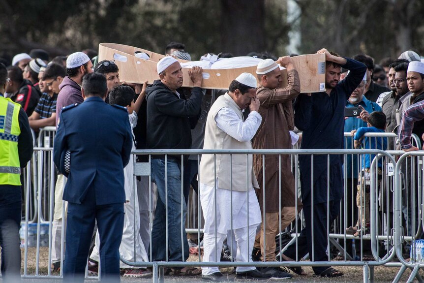 A group carries an open coffin past a crowd while a man appears to wipe tears from his eyes