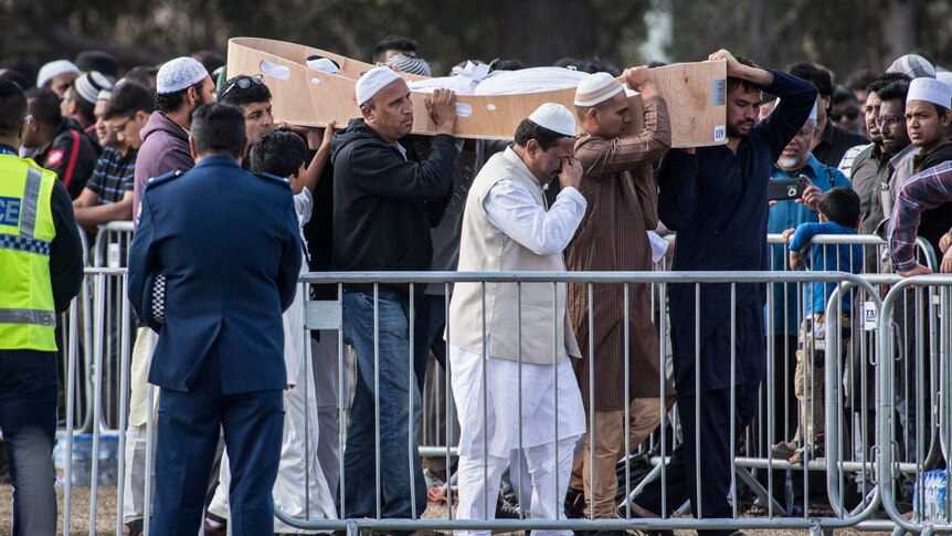 A group carries an open coffin past a crowd while a man appears to wipe tears from his eyes
