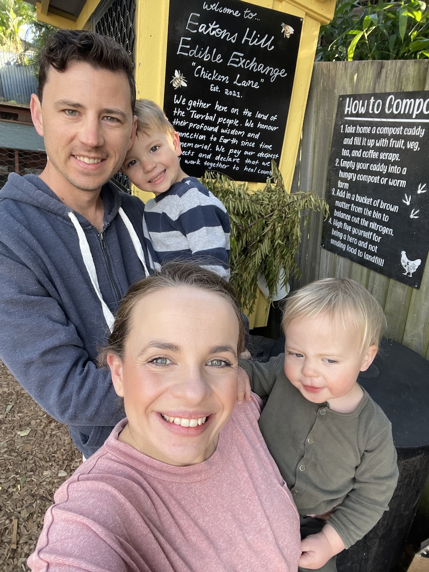 Katie and Tom Irwin carrying their small children in front of yellow hutch with blackboard Eatons Hill Edible Exchange in chalk