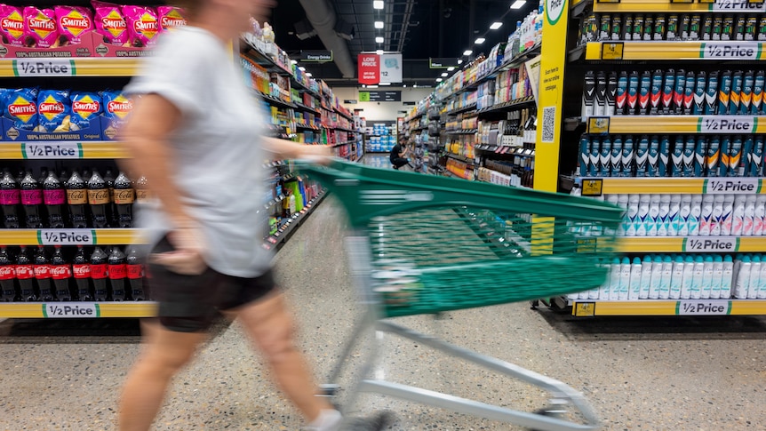 A person wheels their trolley past the end of a supermarket aisle. They are blurred from the motion and are not identifiable.
