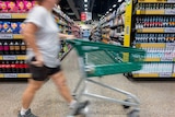 A person wheels their trolley past the end of a supermarket aisle. They are blurred from the motion and are not identifiable.