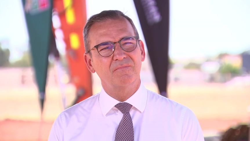 A man wearing a white shirt, purple tie and black glasses stands in front of three coloured flags