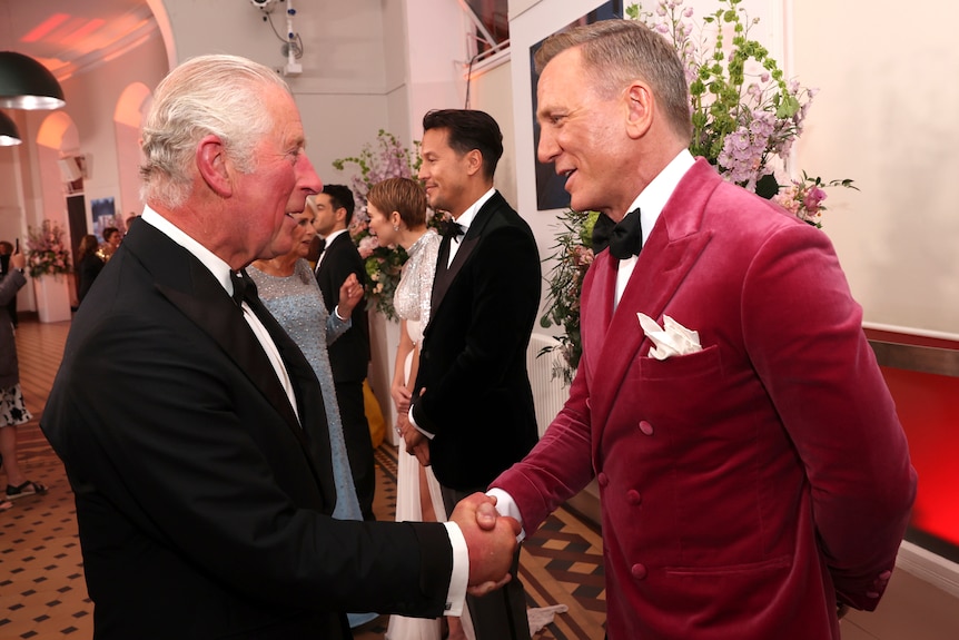 A man in a pink velvet suit shakes the hand of an older man in a black and white suit in a ballroom 