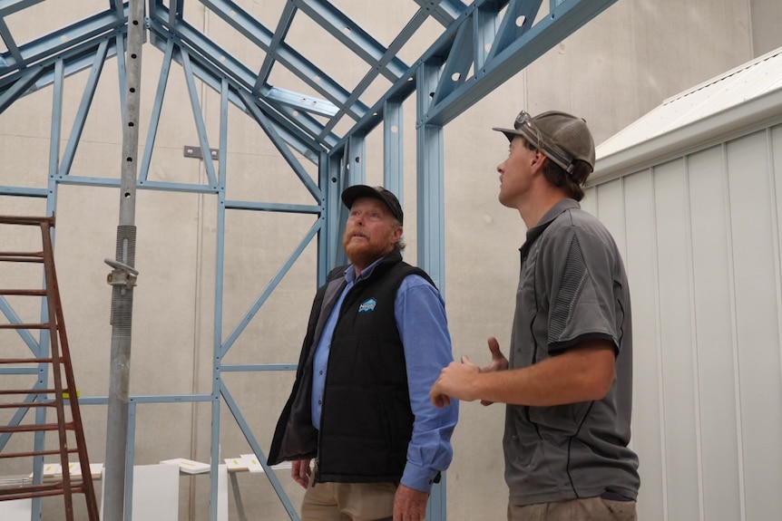 two men in work gear stand in house construction with blue frame looking up
