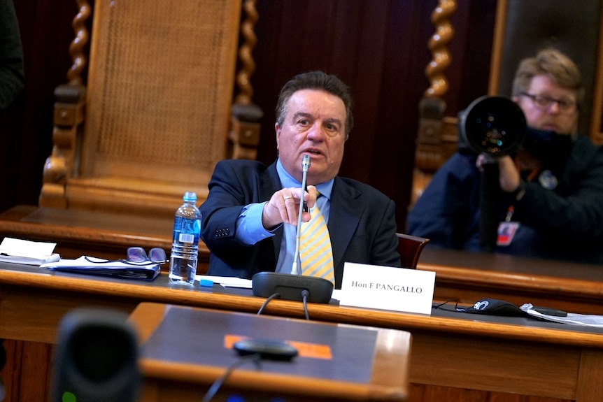 A man sitting behind a microphone points. There is a bottle of water next to him and a name tag