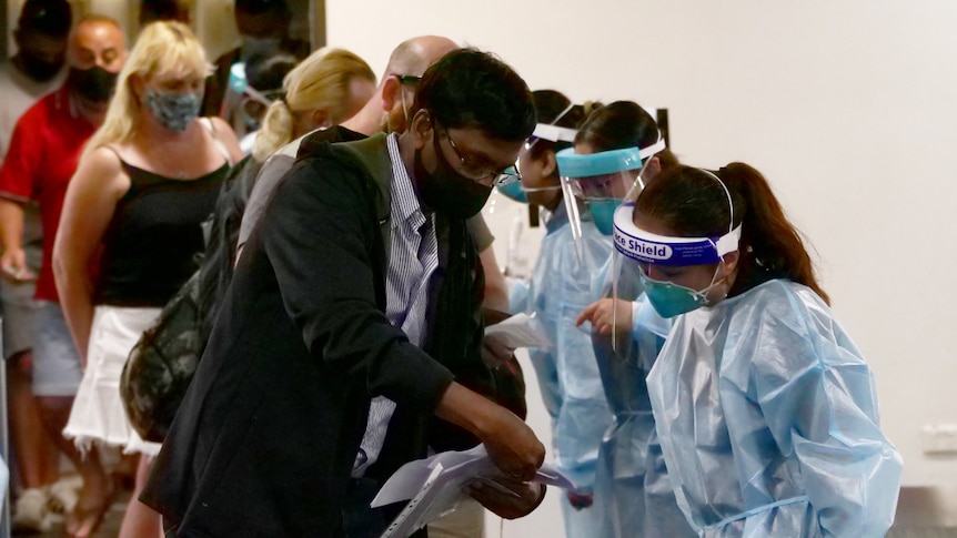 A man shows documentation to a woman in PPE. A queue of people is behind him.