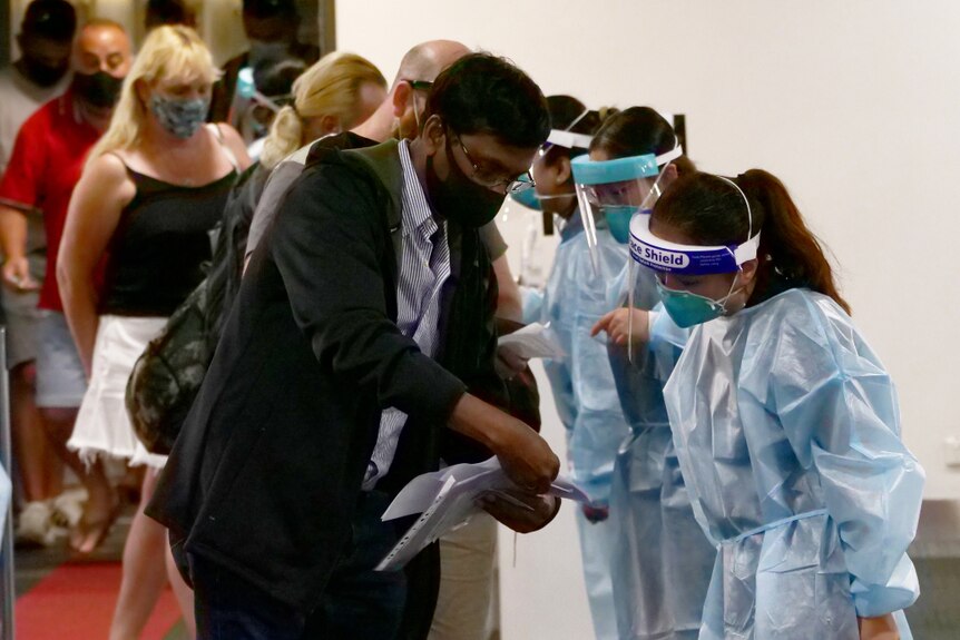 A man shows documentation to a woman in PPE. A queue of people is behind him.