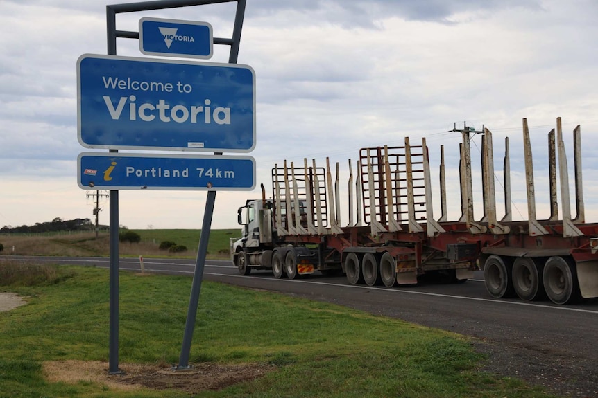A logging truck on the road next to a sign saying "Welcome to Victoria, Portland 74km"