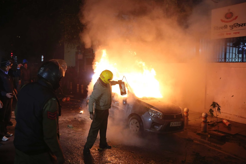 A security officer wearing a helmet tries to put out a fire that is burning in a car on the streets of New Delhi