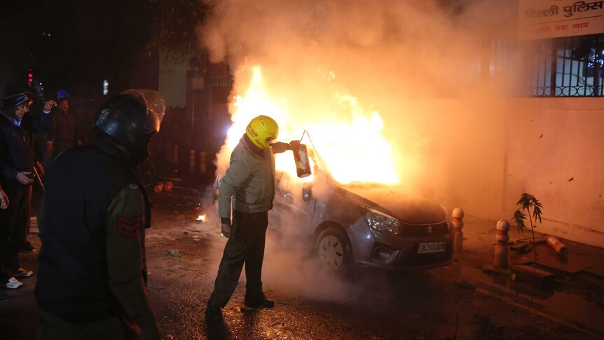 A security officer wearing a helmet tries to put out a fire that is burning in a car on the streets of New Delhi