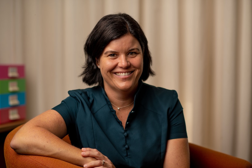 A woman with short, dark brown hair and a warm smile sits on an arm chair.