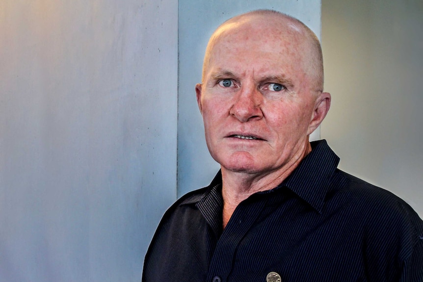 Nearly bald man with blue eyes and a dark blue collared shirt stands in front of bluish grey wall.