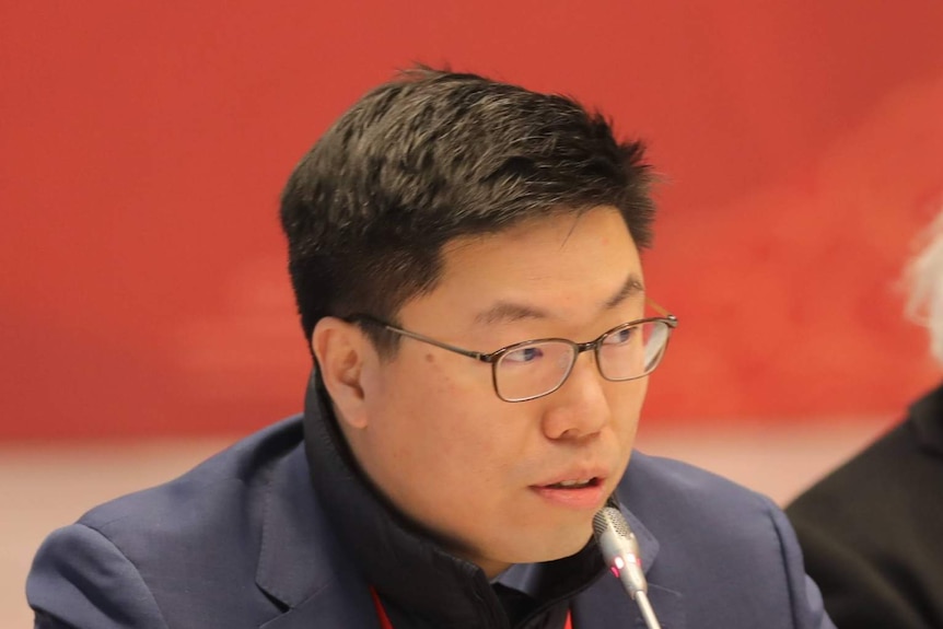 A man with short black hair and glasses sits and speaks into a microphone with a red background.