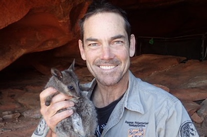 A man wearing a grey safari shirt smiles at the camera while holding a black-footed rock-wallaby