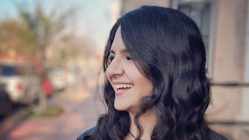 A young woman with long brown hair and black jacket looks to the side smiling broadly. A blurred street is in the background.