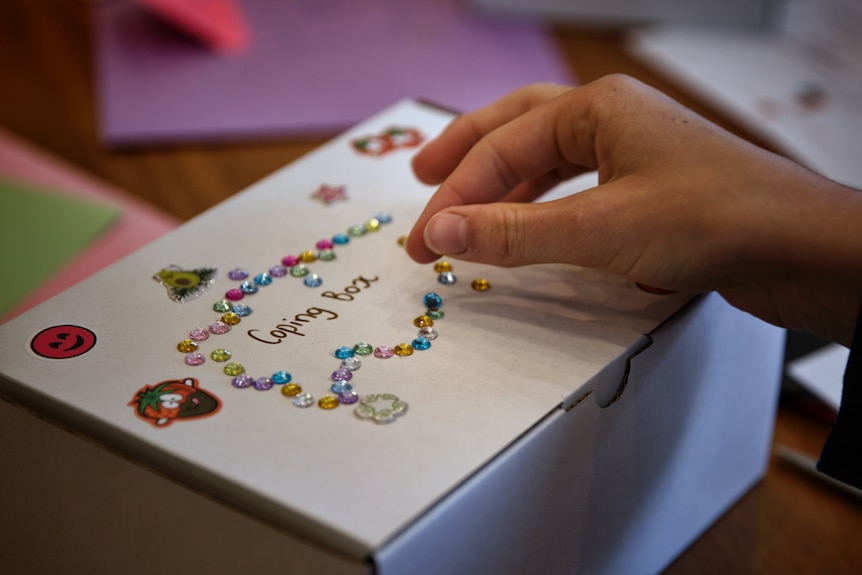 A small white box is decorated with diamontes, stickers of smiley faces, and the words "coping box".