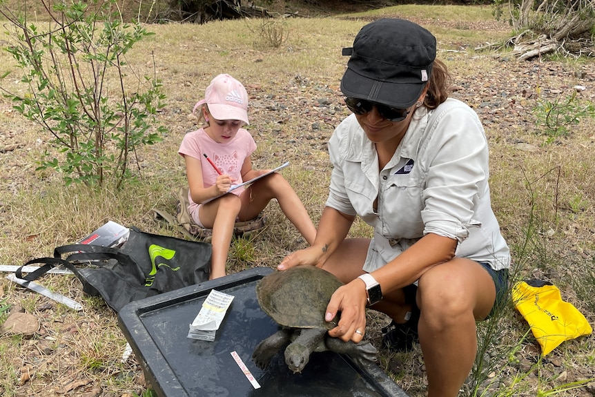 A crouching woman holds a turtle as her young daughter writes down measurements on a pad.