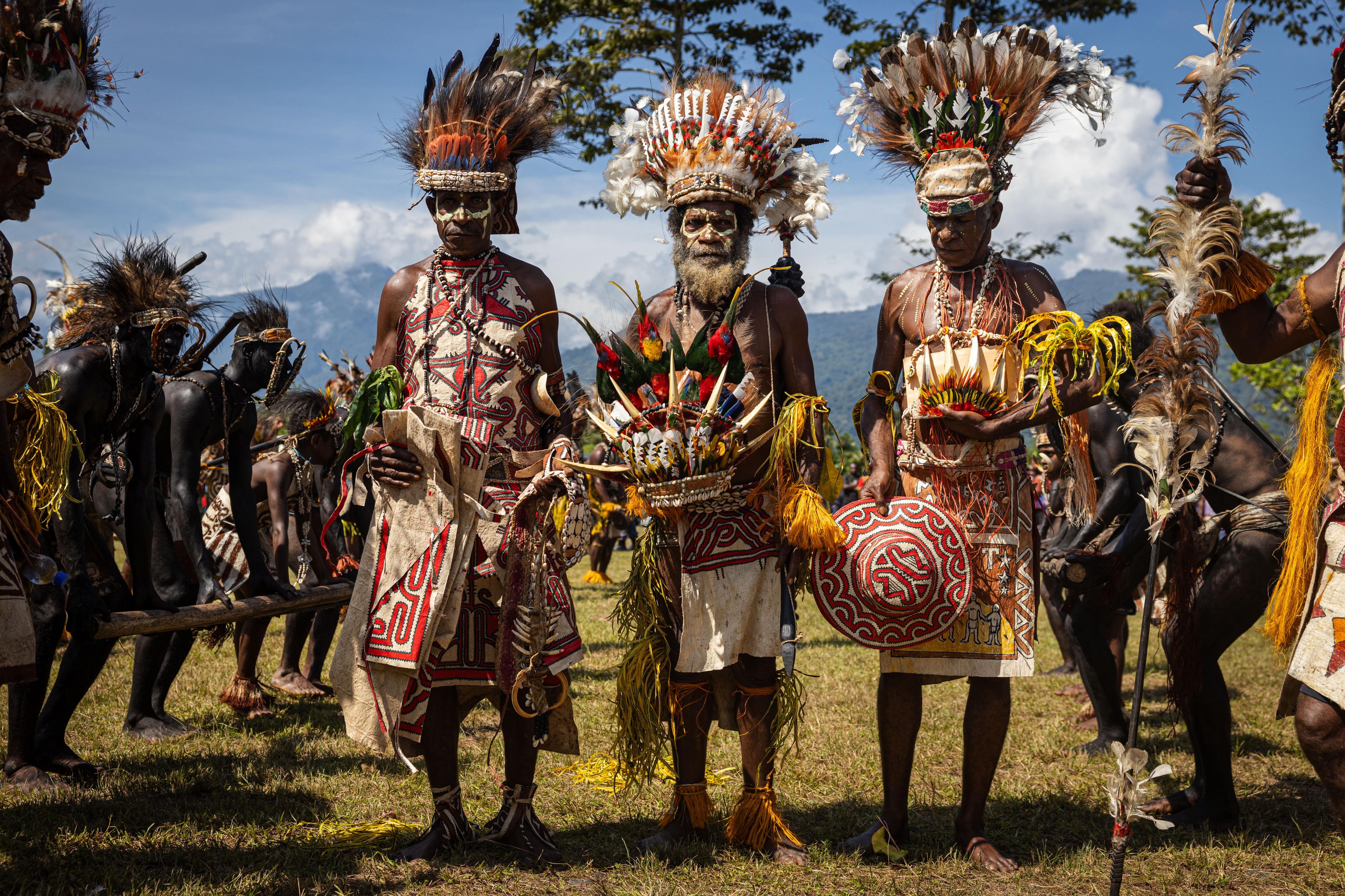 Three men in traditional clothing stand among a crowd holding decorative items, mountains visible behind them.