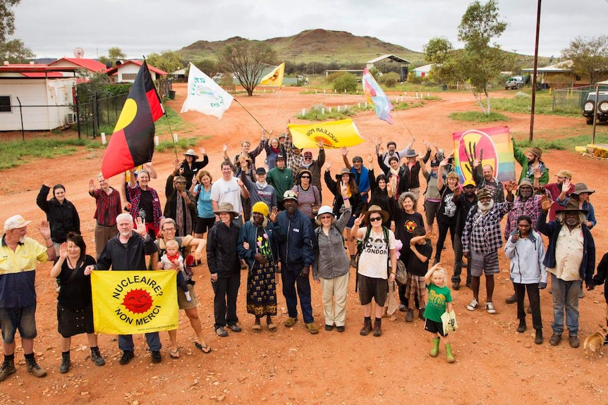 A crowd of Martu people and other protestors wave at the camera holding Aboriginal flags and other banners.