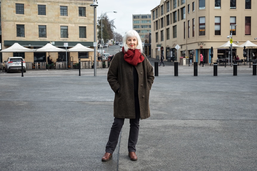 A white woman in her mid 70s with white hair and red scarf and brown winter jacket stands in a street in Tasmania
