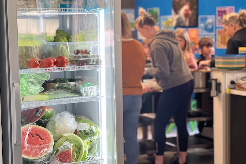 A fridge full of healthy food in a school canteen as children are served lunch in the background.