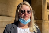 A woman with blonde hair, grey suit jacket and white shirt wearing dark sunglasses and a mask pulled under her chin