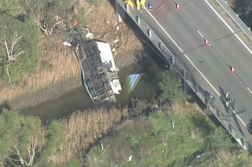 The wreckage of a truck is seen, half in the river, after it crashed off a bridge and emergency service workers gather around.