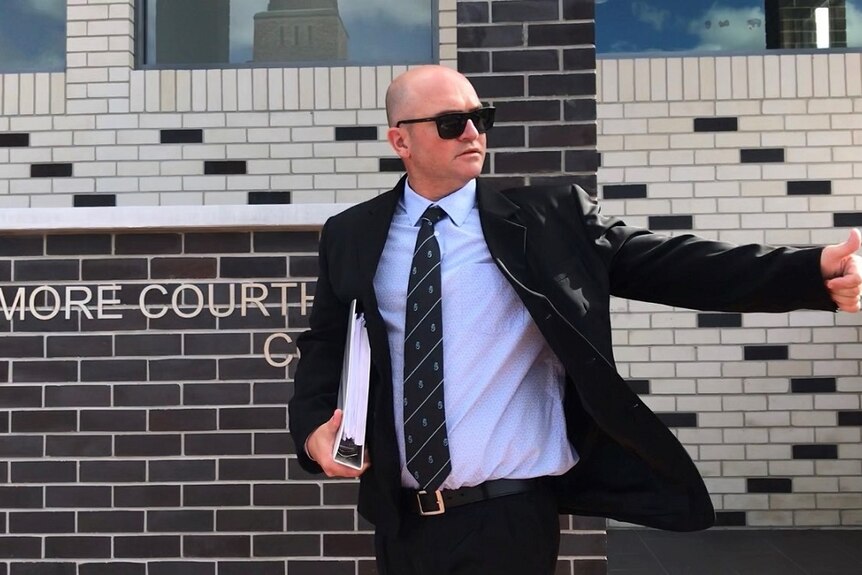 A man with a shaved head, wearing dark sunglasses and a dark suit, leaves a court building.