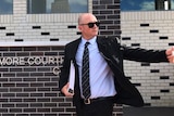 A bald man in a suit and dark sunglasses gestures with his hand as he walks past a court building.