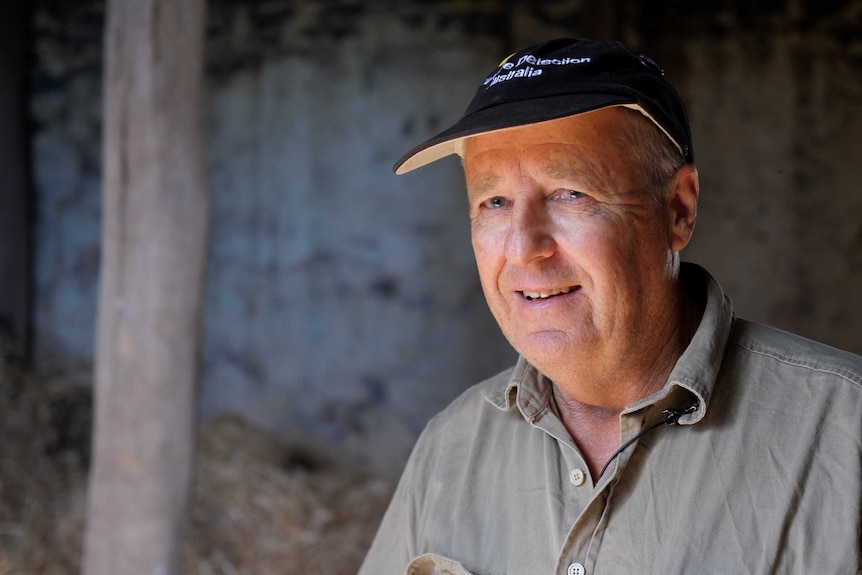 An older man in a cap smiles at the camera in a hay shed