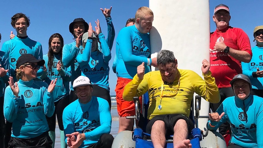 A group of people at the beach, wearing wetsuits and blue vests, cheeringaround a man in a yellow vest sitting in a wheel chair.