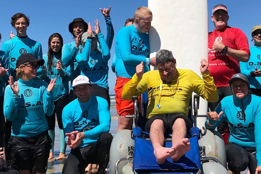 A group of people at the beach, wearing wetsuits and blue vests, cheeringaround a man in a yellow vest sitting in a wheel chair.