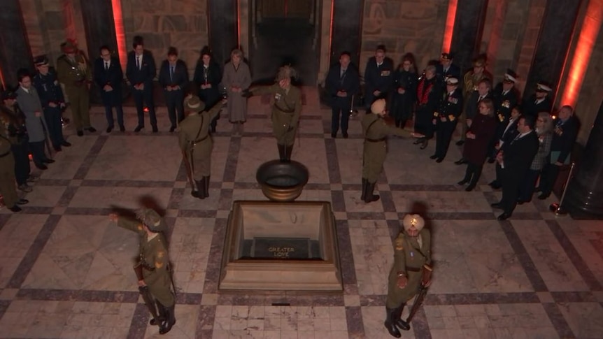 Four soldiers standing positioned at each corner of the stone of Remembrance facing outward. A line of ADF personnel watch on. 