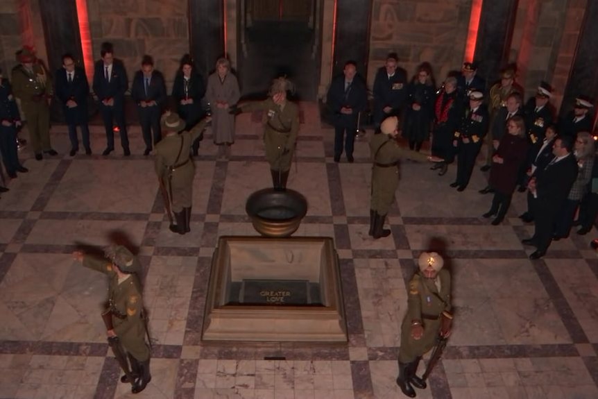 Four soldiers standing positioned at each corner of the stone of Remembrance facing outward. A line of ADF personnel watch on. 